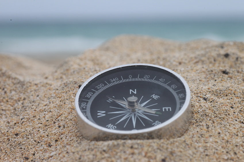 Abgebildet ist ein Kompass, der im Sand am Strand eines Meeres liegt.
