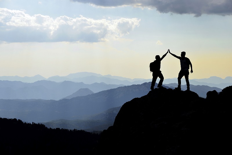 Bild zeigt zwei Wanderer, die einen Berggipfel erreicht haben und sich im Sonnenuntergang abklatschen.