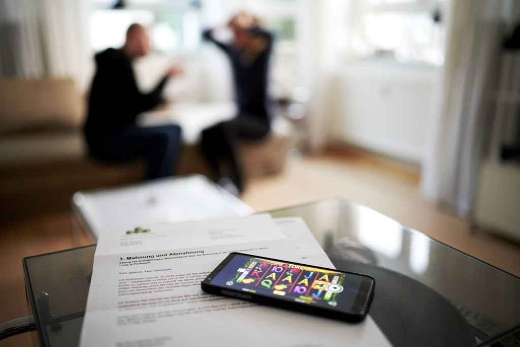 Abgebildet ist im Vordergrun eine MAhnung, wo ein Smartphone mit einer aktivierten Glücksspielseite drauf liegt. Im Hintergrund sitzt ein streitendes Paar.